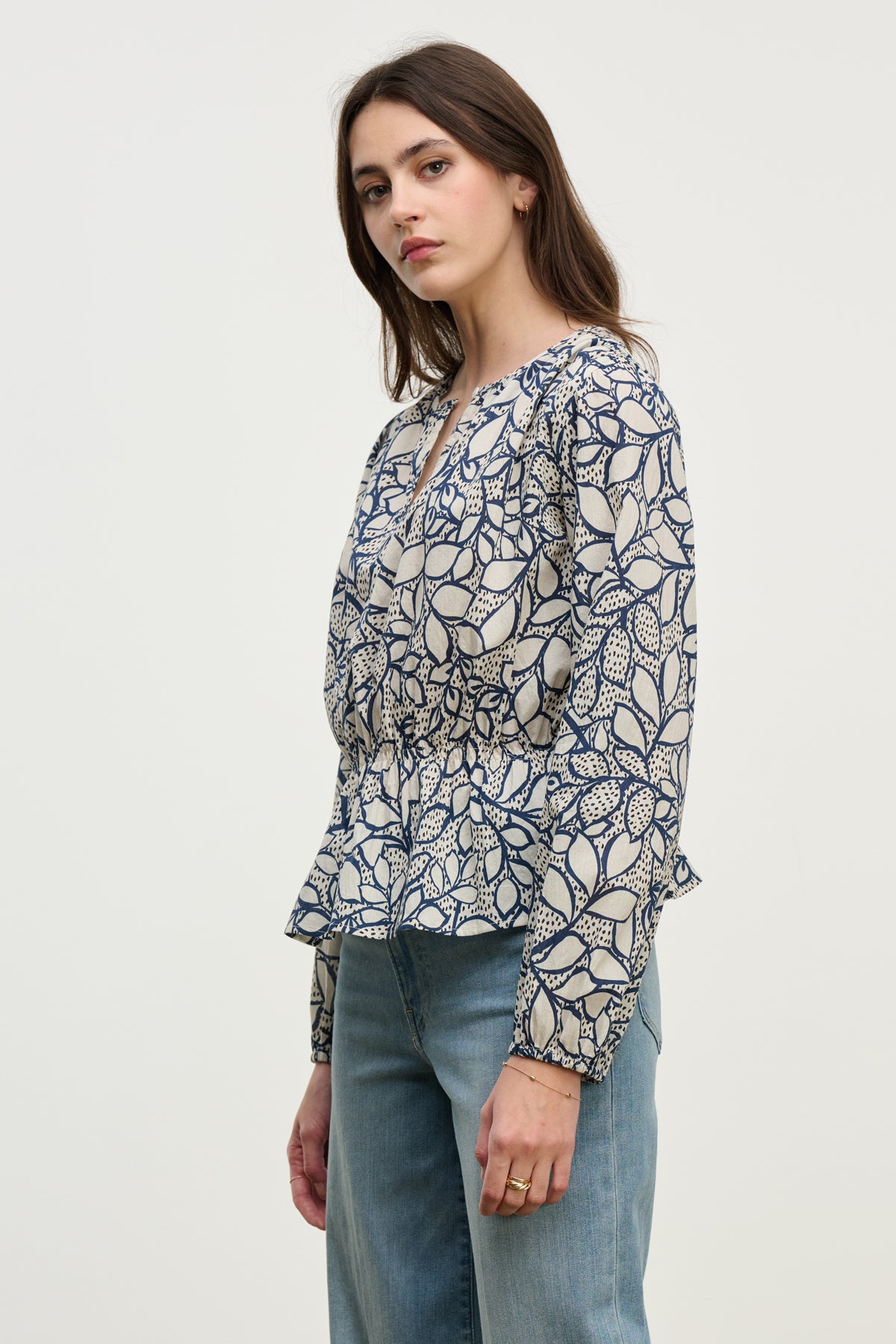A woman wears the WYNNE TOP by Velvet by Graham & Spencer, a long-sleeved patterned cotton voile blouse with a gathered waist and feminine silhouette, paired with blue jeans against a plain light background.-40227172843713