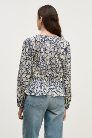 A woman with long brown hair is seen from behind wearing the Velvet by Graham & Spencer WYNNE TOP, a cotton voile blouse with a leaf print, paired with light blue jeans and set against a plain background.