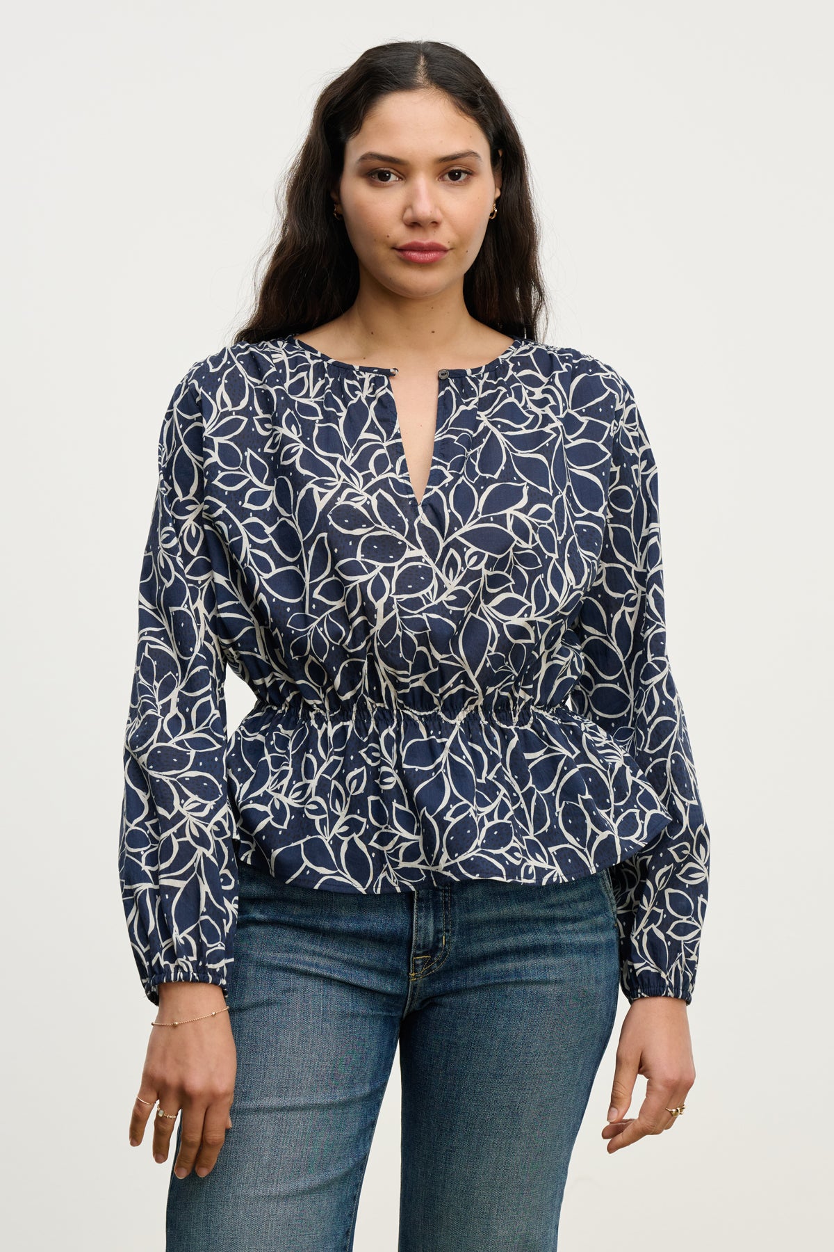 A woman wears the WYNNE TOP by Velvet by Graham & Spencer—a navy cotton voile blouse with a white abstract leaf print and keyhole closure—paired with blue jeans, standing against a plain light background.-40227172909249