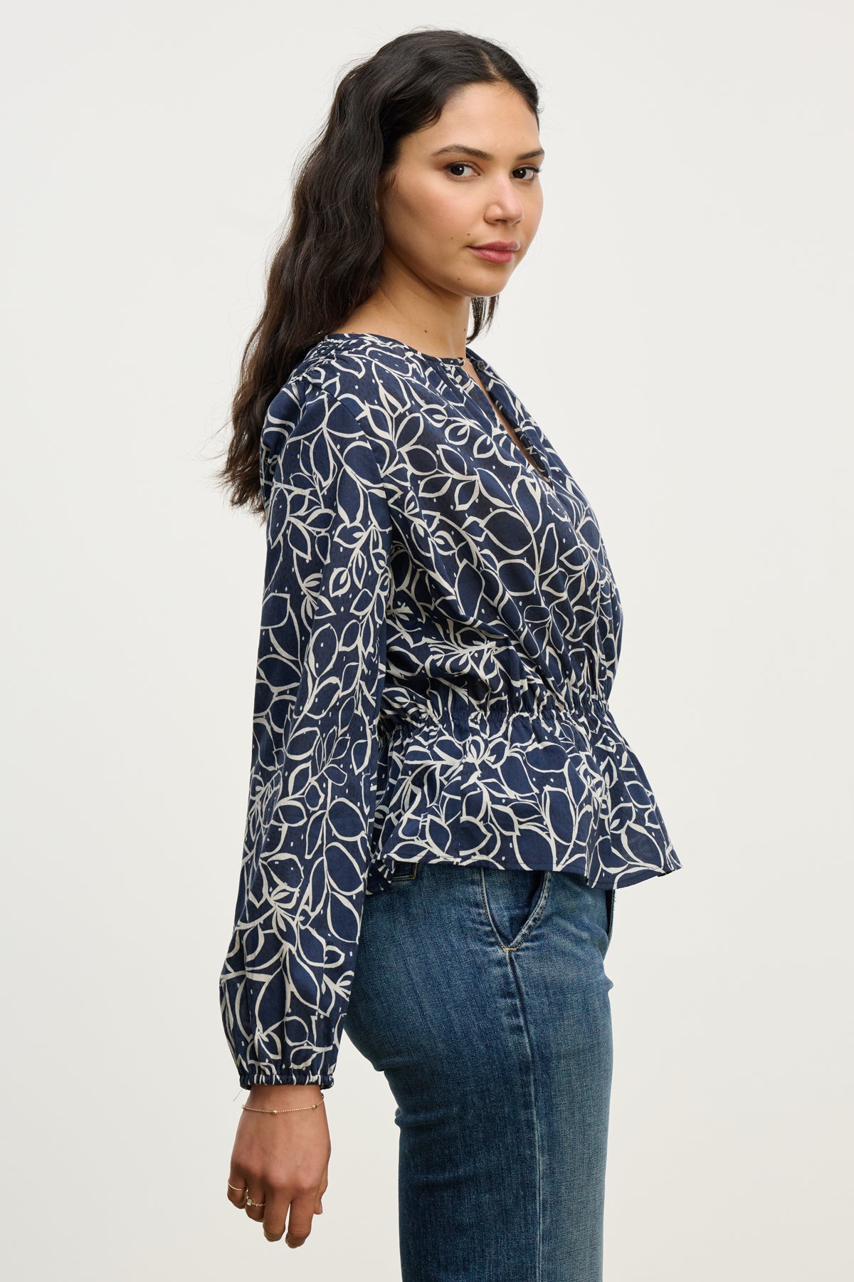 Woman in profile wears the WYNNE TOP by Velvet by Graham & Spencer—a navy blue cotton voile blouse with a white abstract leaf pattern, keyhole closure, and feminine silhouette—paired with blue jeans against a plain light background.-40227172942017