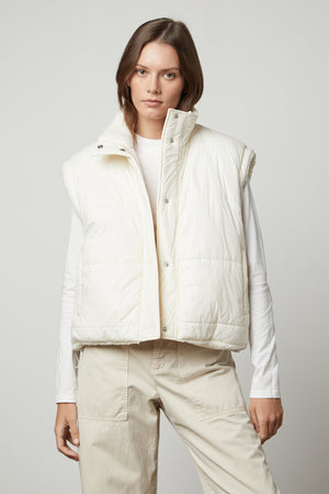 A person is wearing a white ALICIA REVERSIBLE PUFFER SHERPA VEST by Velvet by Graham & Spencer over a white long-sleeve shirt and beige pants. They are standing against a plain gray background.