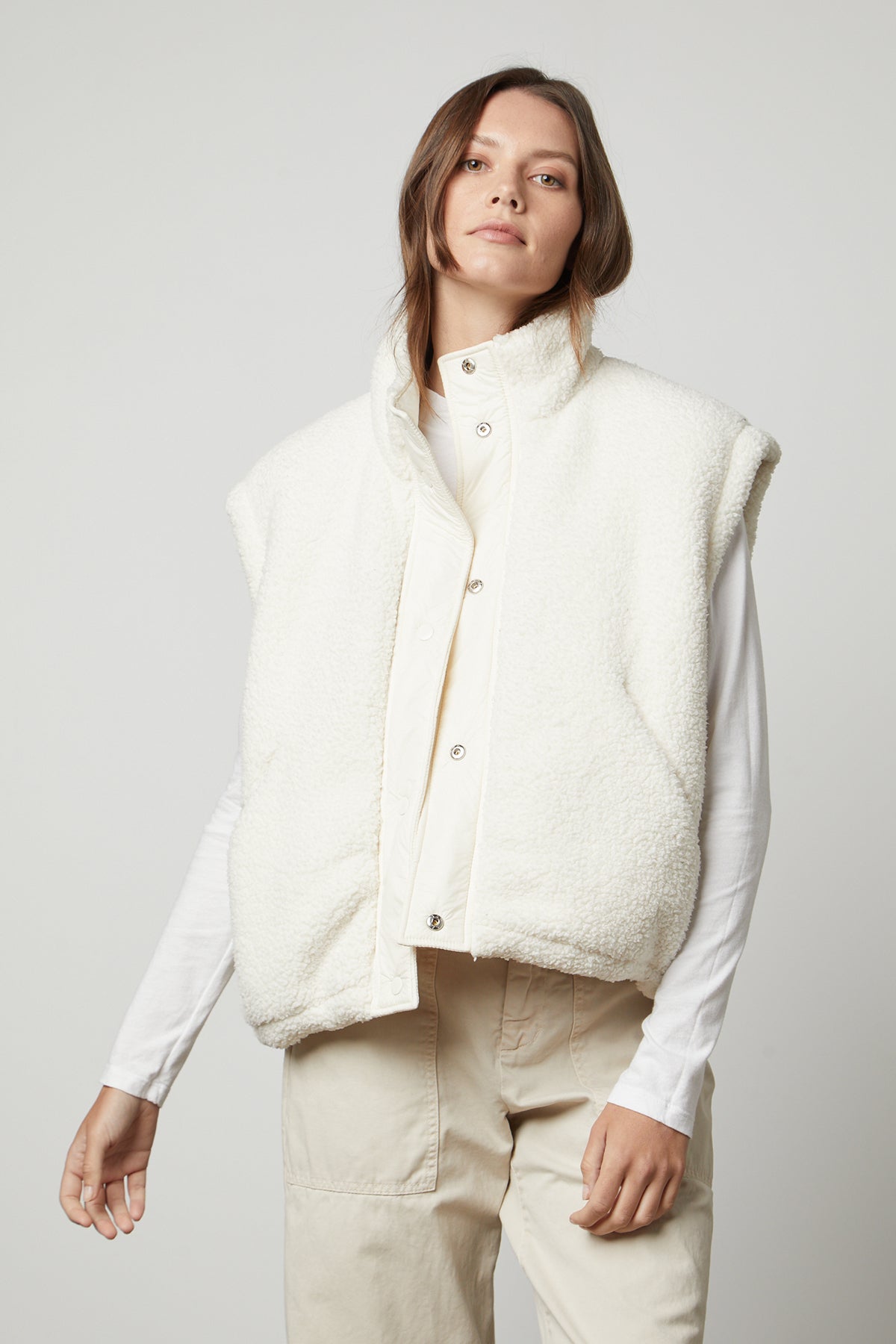   A person stands against a plain background, wearing the ALICIA REVERSIBLE PUFFER SHERPA VEST by Velvet by Graham & Spencer over a long-sleeve white shirt and beige pants, looking directly ahead. 