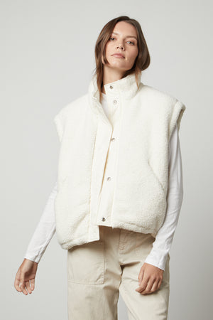 A person stands against a plain background, wearing the ALICIA REVERSIBLE PUFFER SHERPA VEST by Velvet by Graham & Spencer over a long-sleeve white shirt and beige pants, looking directly ahead.