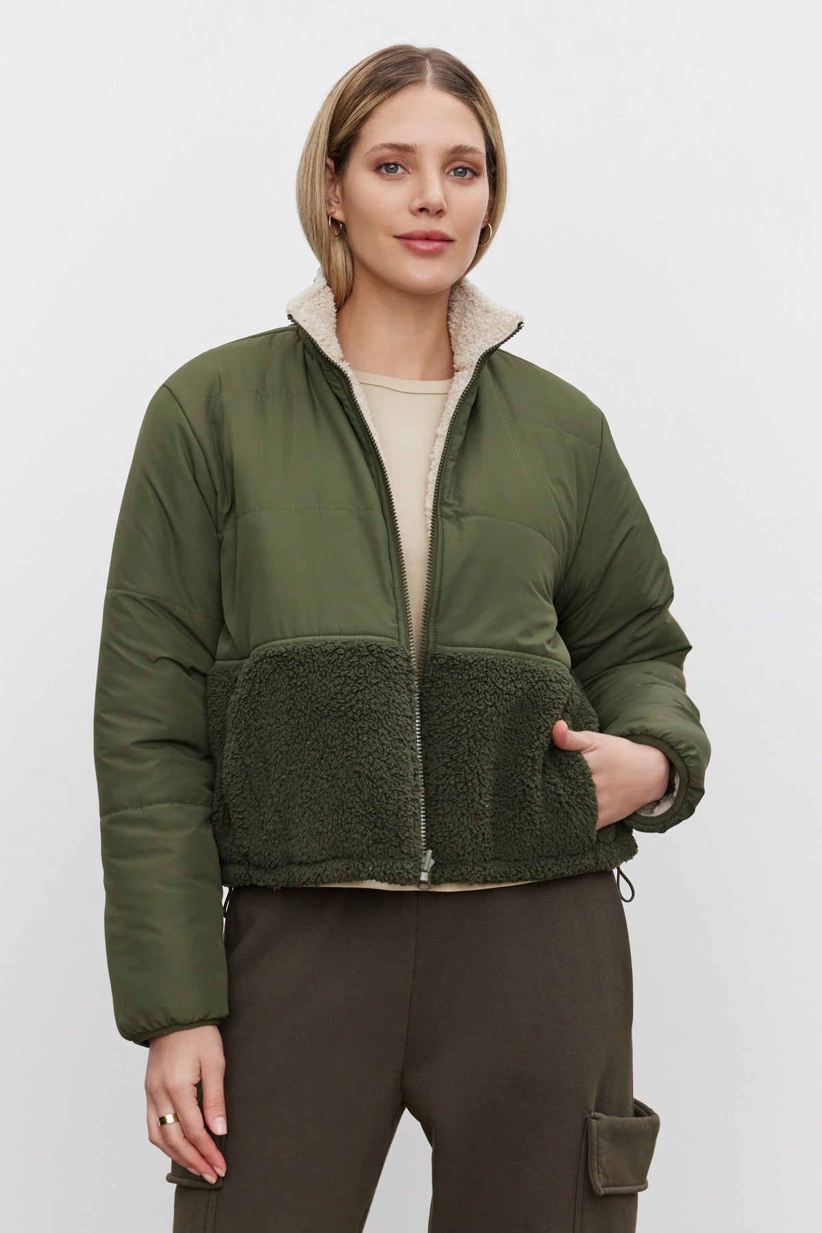   A person with short, blonde hair wearing the Velvet by Graham & Spencer TASHA REVERSIBLE PUFFER SHERPA JACKET, featuring a green quilted durable nylon upper and faux sherpa lower section, along with brown pants, stands against a plain white background. 