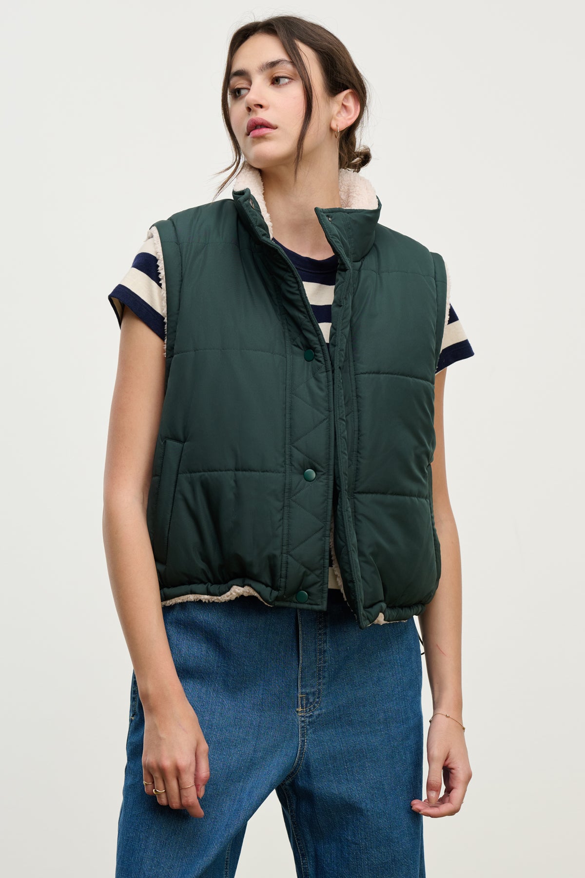 A person models the Velvet by Graham & Spencer CATHY PUFFER SHERPA REVERSIBLE VEST over a striped short-sleeve shirt and blue jeans against a plain light background, highlighting chic transitional layering.-39725322109121