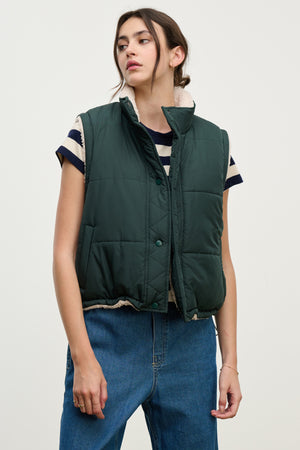 A person models the Velvet by Graham & Spencer CATHY PUFFER SHERPA REVERSIBLE VEST over a striped short-sleeve shirt and blue jeans against a plain light background, highlighting chic transitional layering.