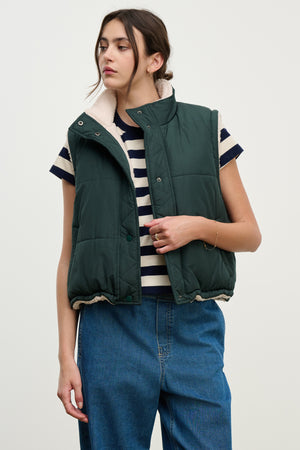 A person wears the Velvet by Graham & Spencer CATHY PUFFER SHERPA REVERSIBLE VEST over a black-and-white striped tee and blue jeans, standing against a plain light background to showcase stylish transitional layering.