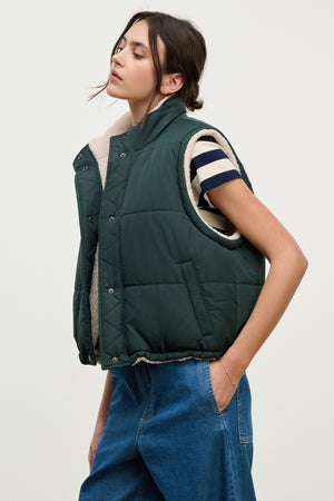 A woman models the CATHY PUFFER SHERPA REVERSIBLE VEST by Velvet by Graham & Spencer over a striped tee and blue jeans, showcasing chic transitional layering with one hand in her pocket against a plain background.