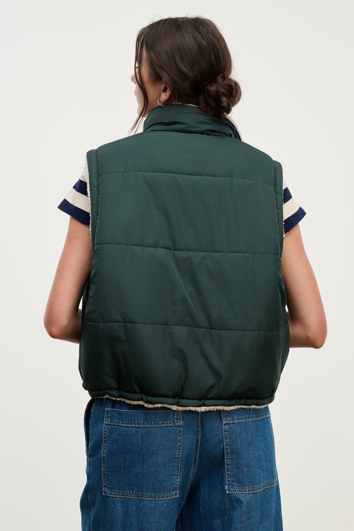 A person stands with their back to the camera, showcasing transitional layering with the Velvet by Graham & Spencer CATHY PUFFER SHERPA REVERSIBLE VEST in dark green, worn over a striped short-sleeve shirt and blue jeans.-39725322010817