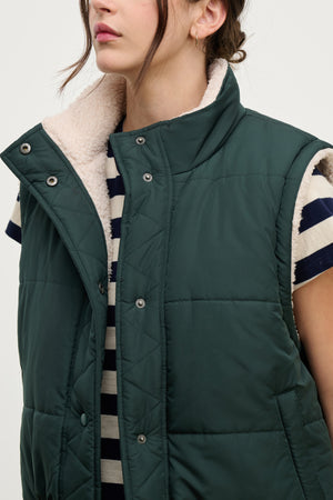 A person wears the Velvet by Graham & Spencer CATHY Puffer Sherpa Reversible Vest over a black and white striped shirt. The cozy vest, ideal for transitional layering, is unbuttoned. The person has shoulder-length brown hair.