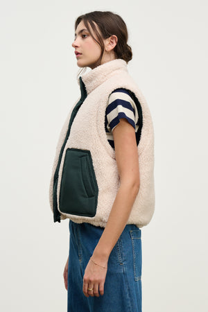 A woman in profile wears the Velvet by Graham & Spencer CATHY PUFFER SHERPA REVERSIBLE VEST in beige with dark green pockets over a striped tee and blue jeans—ideal for transitional layering—set against a plain light background.