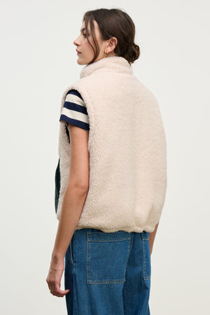 A person stands facing away, wearing the CATHY PUFFER SHERPA REVERSIBLE VEST by Velvet by Graham & Spencer over a black and white striped shirt and blue jeans, ideal for transitional layering against a plain light background.
