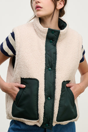 A person wears the CATHY PUFFER SHERPA REVERSIBLE VEST by Velvet by Graham & Spencer, featuring cream faux sherpa with dark green quilted snaps and pockets, layered over a navy and white striped shirt—ideal for transitional weather.