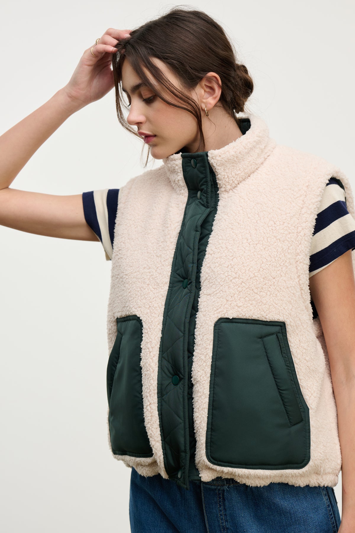 A woman models the Velvet by Graham & Spencer CATHY PUFFER SHERPA REVERSIBLE VEST, featuring dark green quilted pockets over a striped short-sleeve shirt, ideal for transitional layering, against a simple light backdrop.-39725320503489