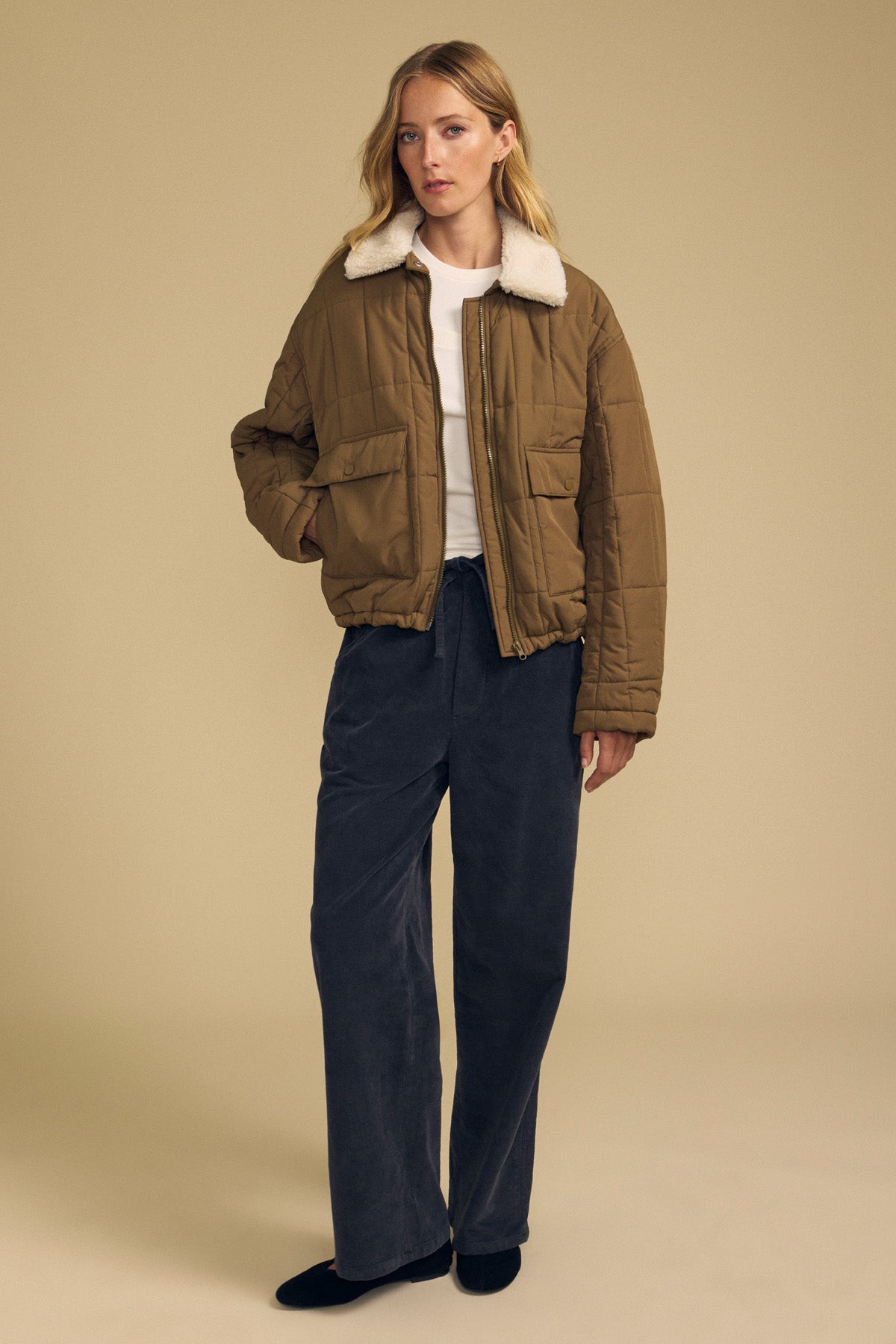 A woman stands against a beige background wearing the NOA QUILTED JACKET by Velvet by Graham & Spencer, featuring a brown quilted design with a faux sherpa collar, paired with a white shirt, wide-leg dark pants, and black shoes.