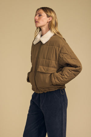A person wearing the NOA QUILTED JACKET by Velvet by Graham & Spencer—a brown quilted jacket featuring lightweight insulation and a white faux sherpa collar—paired with navy corduroy pants, stands sideways against a beige background.