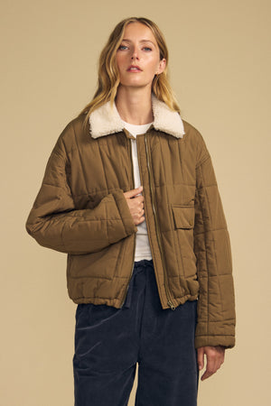 A woman models the Velvet by Graham & Spencer NOA QUILTED JACKET—featuring lightweight insulation and a cozy faux sherpa collar—styled with a white t-shirt and navy blue pants against a plain beige background.