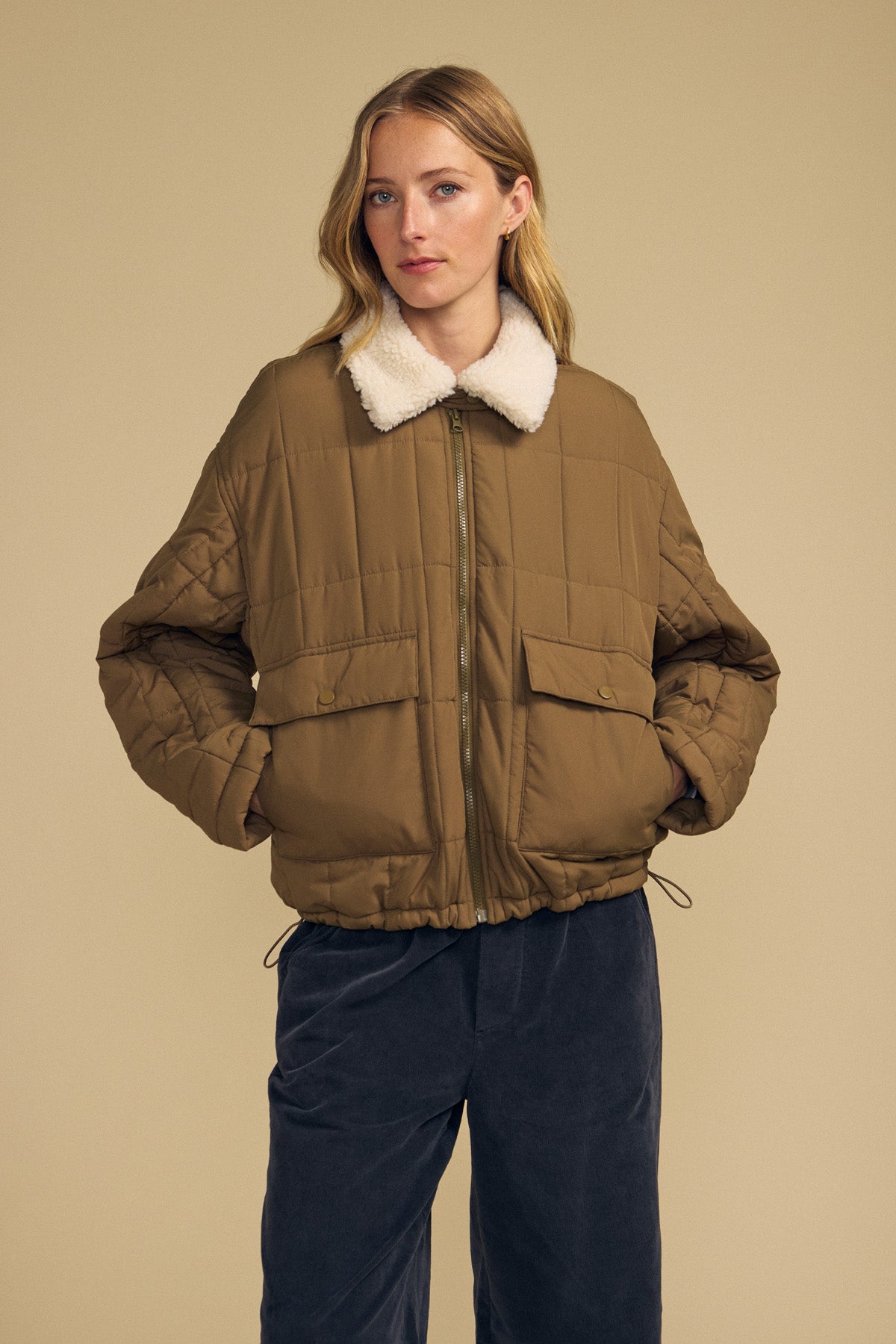 A woman wearing the Velvet by Graham & Spencer NOA Quilted Jacket, featuring lightweight insulation and a white faux sherpa collar, stands against a beige background in dark pants.-41964703285441