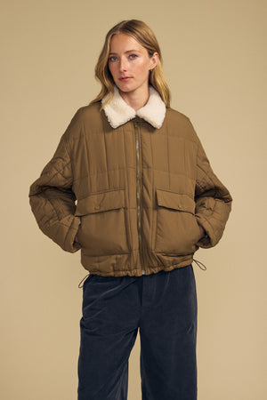 A woman wearing the Velvet by Graham & Spencer NOA Quilted Jacket, featuring lightweight insulation and a white faux sherpa collar, stands against a beige background in dark pants.