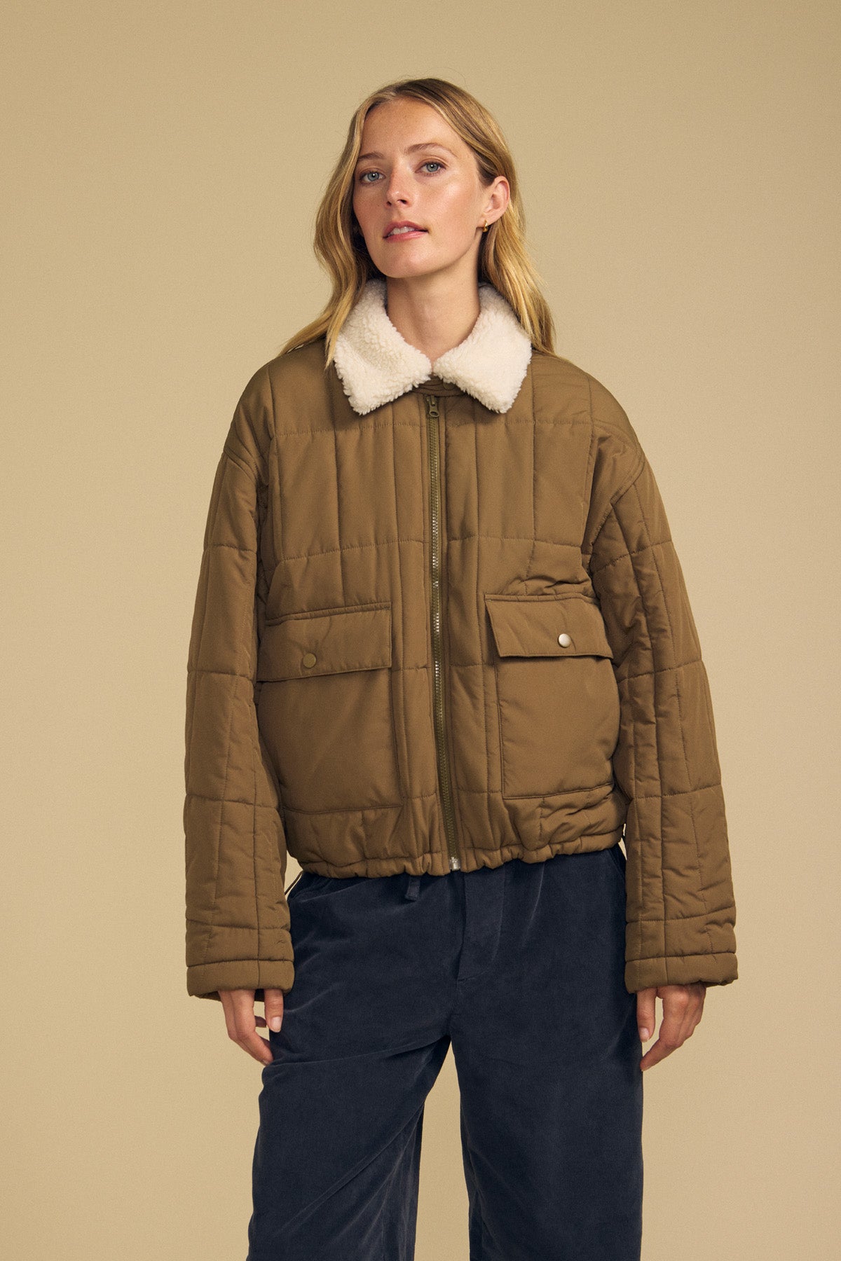 A woman wearing the NOA QUILTED JACKET by Velvet by Graham & Spencer—featuring a tan quilted design with lightweight insulation and a faux sherpa collar—is paired with navy pants and stands against a plain beige background.
