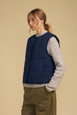 Wearing the ONYX REVERSIBLE VEST by Velvet by Graham & Spencer—a navy quilted vest with faux sherpa lining—over a beige sweater and olive green pants, a woman stands against a plain beige background, gazing to the side.