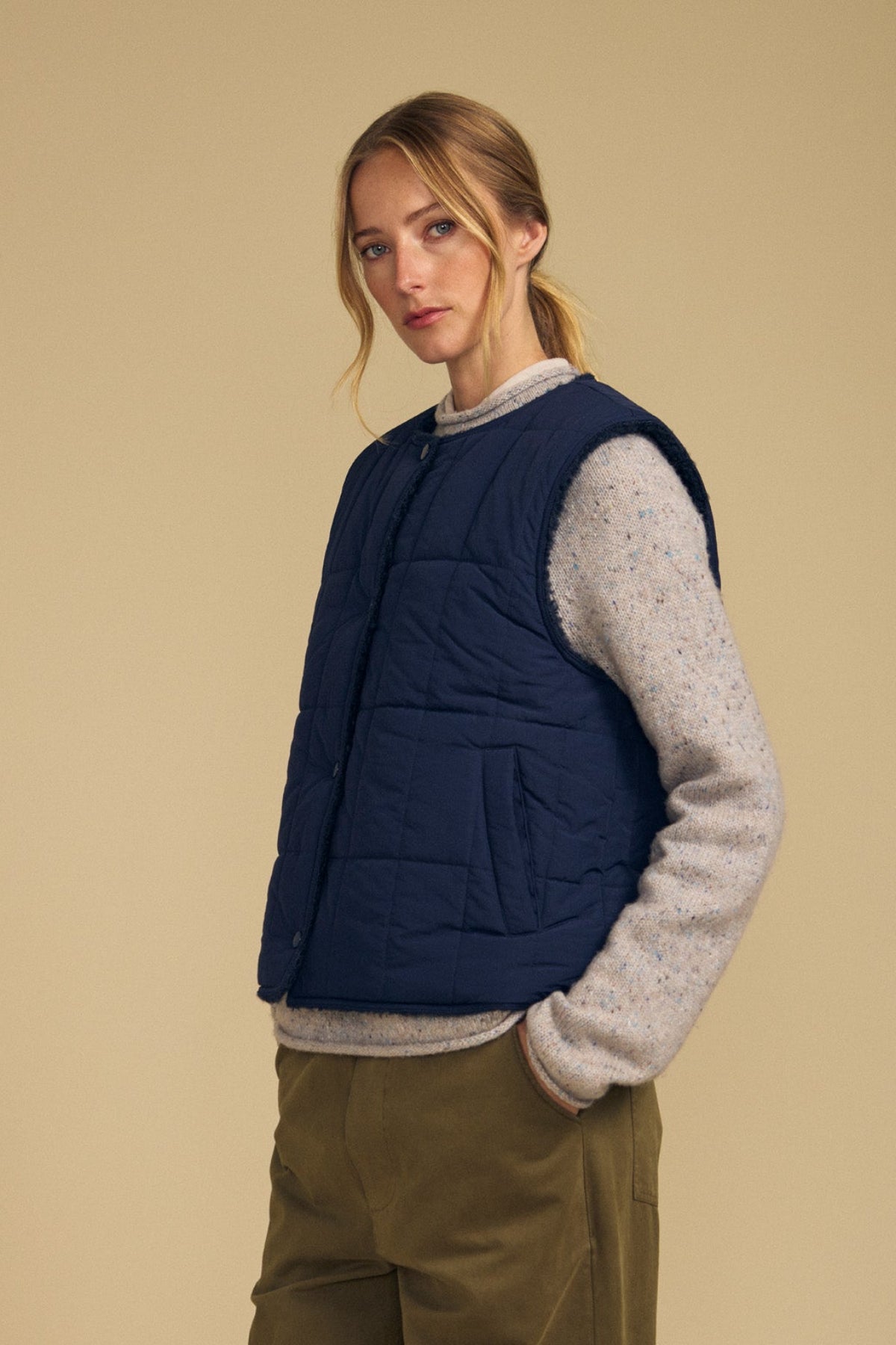 Wearing the ONYX REVERSIBLE VEST by Velvet by Graham & Spencer over a light gray sweater and olive pants, a person stands against a beige background, looking at the camera.