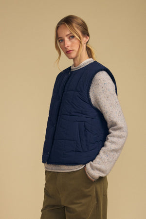 Wearing the ONYX REVERSIBLE VEST by Velvet by Graham & Spencer over a light gray sweater and olive pants, a person stands against a beige background, looking at the camera.