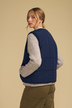 A person with blond hair, wearing a gray sweater, olive green pants, and the ONYX REVERSIBLE VEST by Velvet by Graham & Spencer in navy quilted fabric, stands facing away and looks over their shoulder against a beige background.