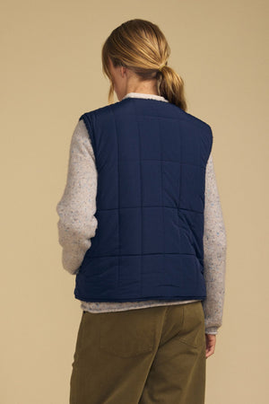 A person with light brown hair in a low ponytail wears the ONYX REVERSIBLE VEST by Velvet by Graham & Spencer—a navy quilted, reversible vest—layered over a light sweater and paired with olive green pants, standing with their back to the camera against a beige background.