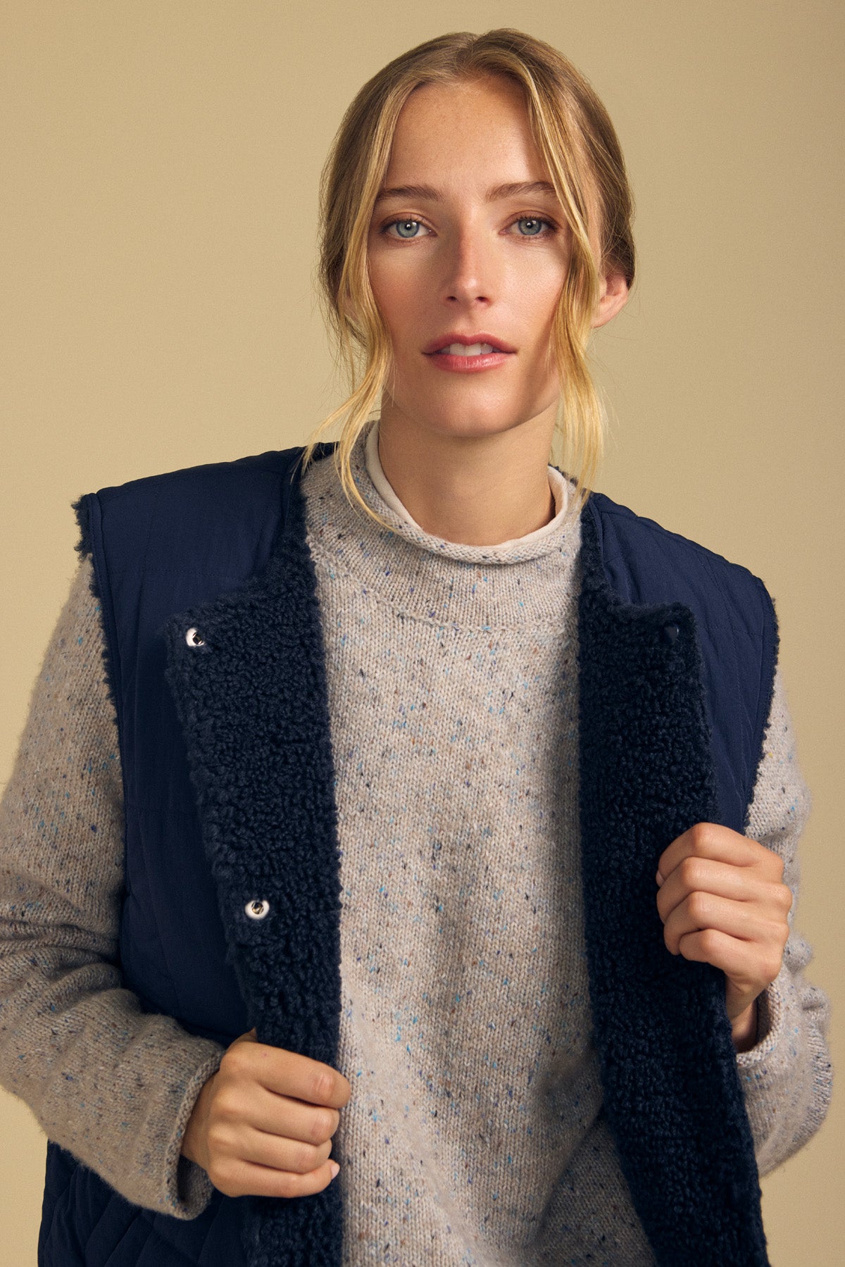 A woman with blonde hair wears the ONYX REVERSIBLE VEST by Velvet by Graham & Spencer over a light gray sweater as she looks at the camera against a beige background.-41964704497857