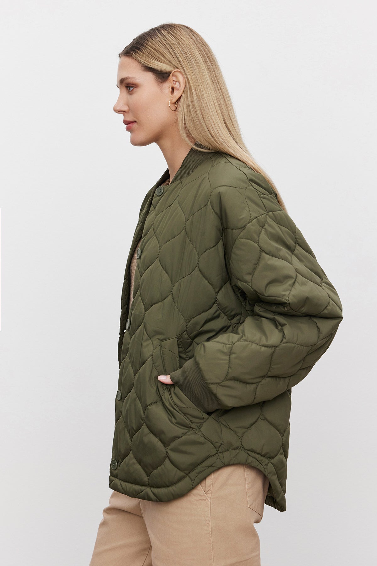   A person with long blonde hair wears an oversized CARIE QUILTED JACKET by Velvet by Graham & Spencer, featuring a scooped hemline and beige pants, standing in profile against a plain white background. 