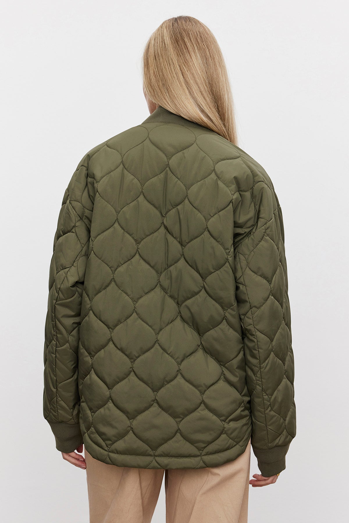   A person with long blonde hair is facing away, showcasing the Velvet by Graham & Spencer CARIE QUILTED JACKET in olive green with a scooped hemline, paired with beige pants against a plain white background. 