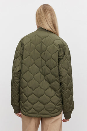 A person with long blonde hair is facing away, showcasing the Velvet by Graham & Spencer CARIE QUILTED JACKET in olive green with a scooped hemline, paired with beige pants against a plain white background.