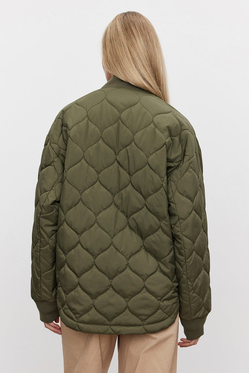 A person with long blonde hair is facing away, showcasing the Velvet by Graham & Spencer CARIE QUILTED JACKET in olive green with a scooped hemline, paired with beige pants against a plain white background.