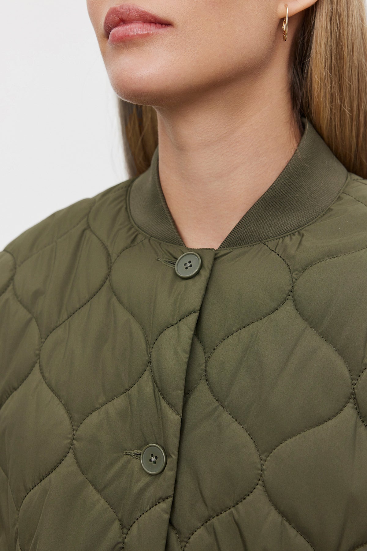   Close-up of a person wearing the Velvet by Graham & Spencer CARIE QUILTED JACKET in olive-green, featuring button details. The oversized fit lends a cozy look, accentuated by a small gold hoop earring. The person's long, blonde hair cascades down, with only the lower part of their face visible. 