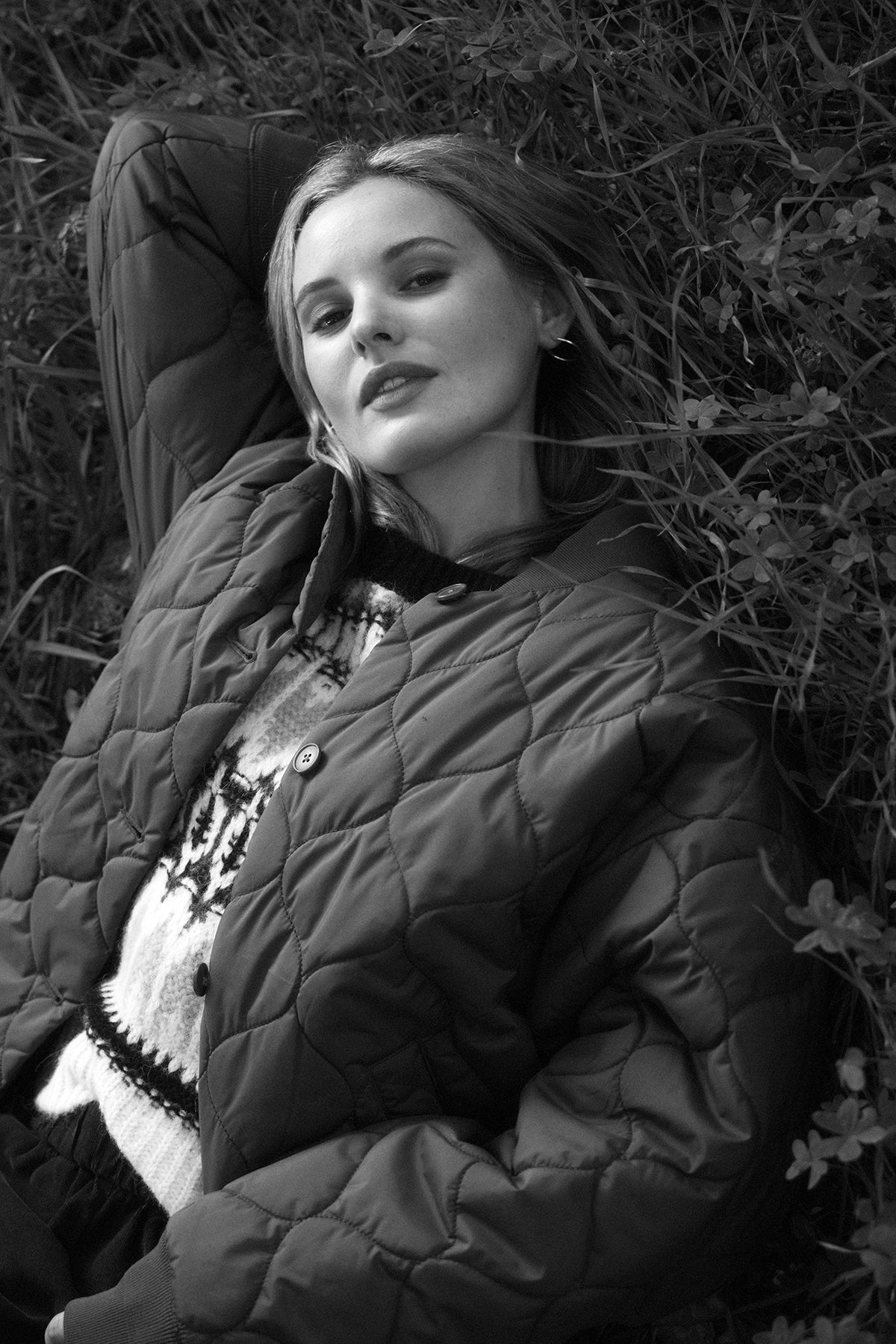   Black and white image of a person in an oversized CARIE QUILTED JACKET by Velvet by Graham & Spencer and sweater, lying down on grass, looking at the camera with one arm resting behind their head. 