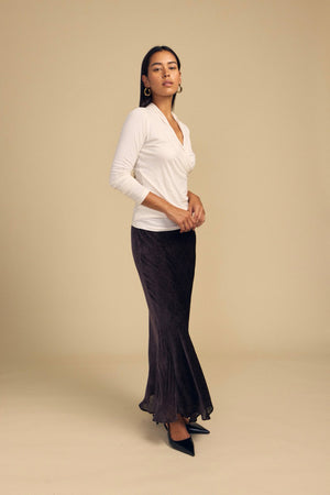 A model wears a white long-sleeved wrap top and a dark, bias-cut QUINN DEVORE DOT SKIRT with a subtle burnout texture, paired with black pointed-toe heels.