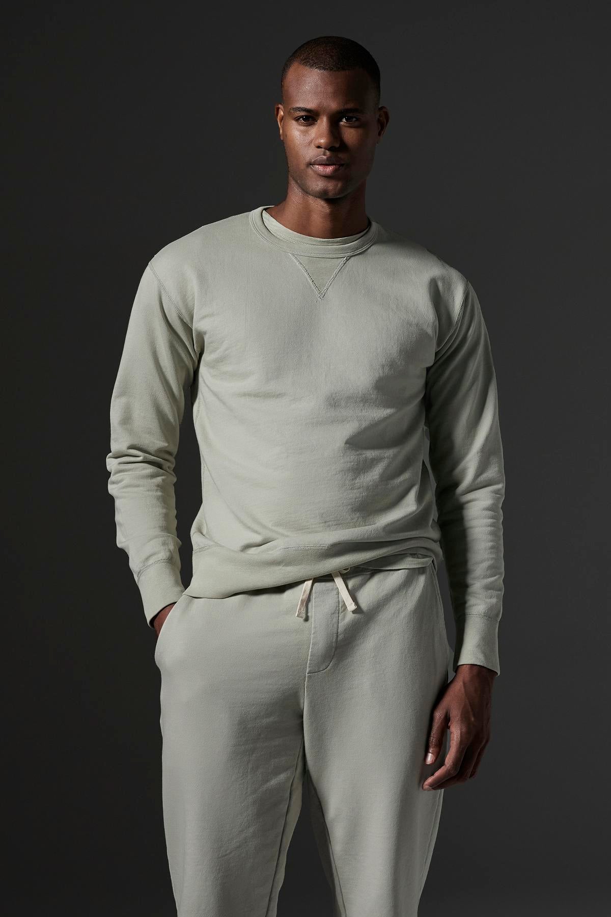   A person wears a light gray sweatshirt and Velvet by Graham & Spencer DUSTY SWEATPANT, featuring an elastic drawstring waist and ribbed cuffs, standing with one hand in their pocket against a dark background. 