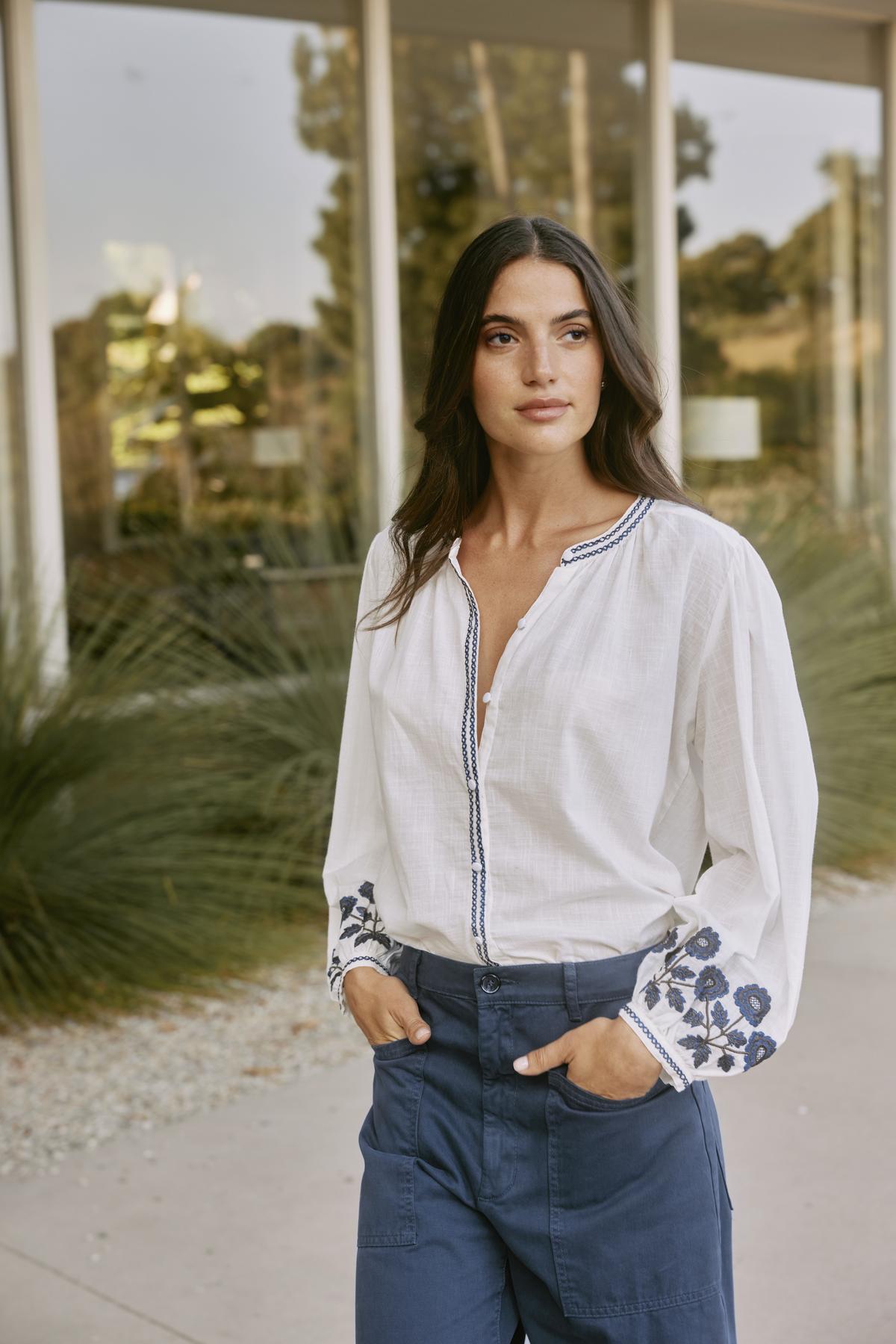 A woman wearing the RAHWA TOP by Velvet by Graham & Spencer stands outdoors near a building with large windows and lush plants. The lightweight cotton blouse, featuring floral embroidery and a button front, elegantly catches the breeze.-38512732733633