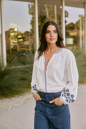 A woman wearing the RAHWA TOP by Velvet by Graham & Spencer stands outdoors near a building with large windows and lush plants. The lightweight cotton blouse, featuring floral embroidery and a button front, elegantly catches the breeze.