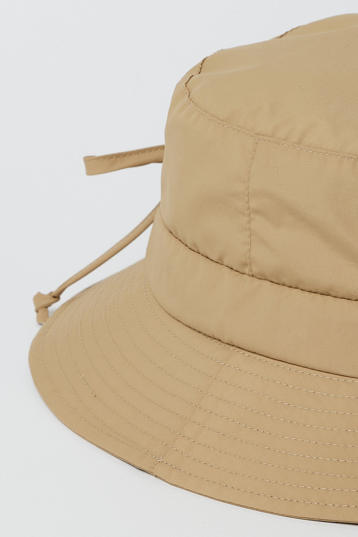Close-up of the RAIN HAT by Velvet by Graham & Spencer, a light brown, water-resistant 100% nylon bucket hat with visible stitching and an adjustable back drawstring, set against a plain white background.-39301129568449
