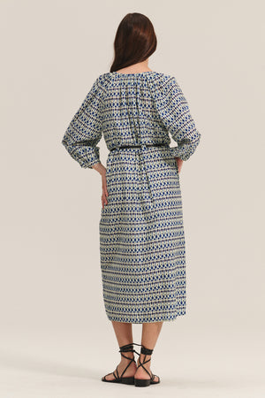 A woman faces away, wearing the RAINE DRESS by Velvet by Graham & Spencer— a silk cotton voile midi with pleated details and a belted waist—styled with black strappy sandals.