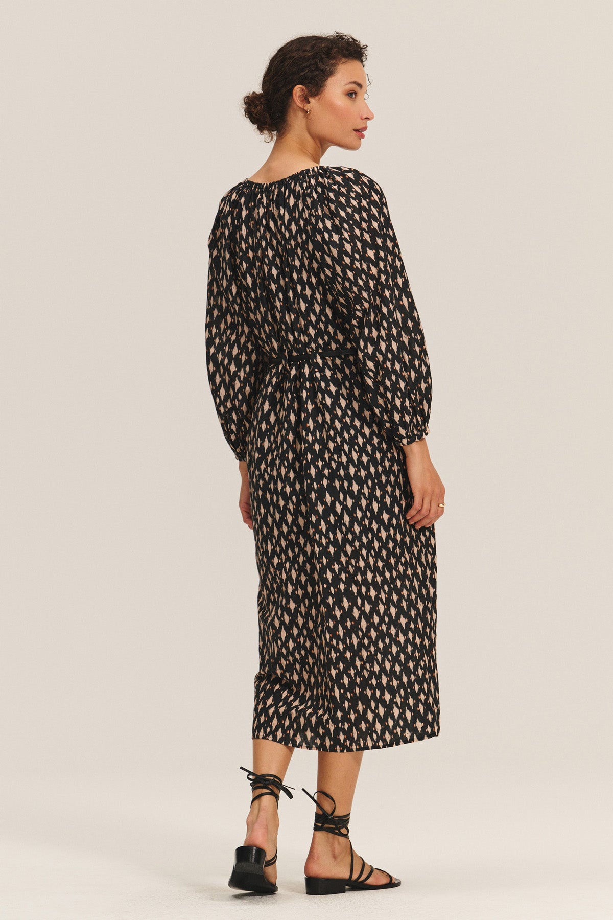 A woman stands with her back turned, wearing the RAINE DRESS by Velvet by Graham & Spencer—a black silk cotton voile midi dress with a beige geometric pattern, pleated details, and black strappy sandals.-40688718250177
