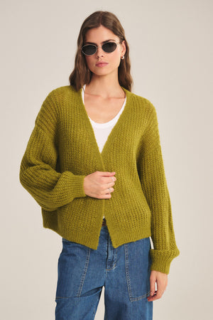 A woman in sunglasses, a white shirt, blue jeans, and the RANYA ALPACA CARDIGAN by Velvet by Graham & Spencer stands against a plain background, highlighting cozy layering.