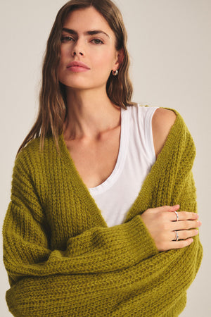 A woman with long brown hair wears the RANYA ALPACA CARDIGAN by Velvet by Graham & Spencer in textured olive green over a white shirt, looking at the camera with a neutral expression—ideal for effortless layering.