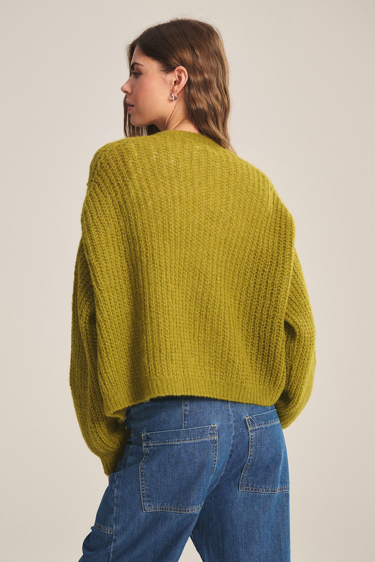   A person with wavy brown hair is seen from the back in a yellow-green Velvet by Graham & Spencer RANYA ALPACA CARDIGAN and blue jeans, set against a plain light background. 