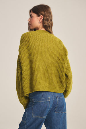 A person with wavy brown hair is seen from the back in a yellow-green Velvet by Graham & Spencer RANYA ALPACA CARDIGAN and blue jeans, set against a plain light background.
