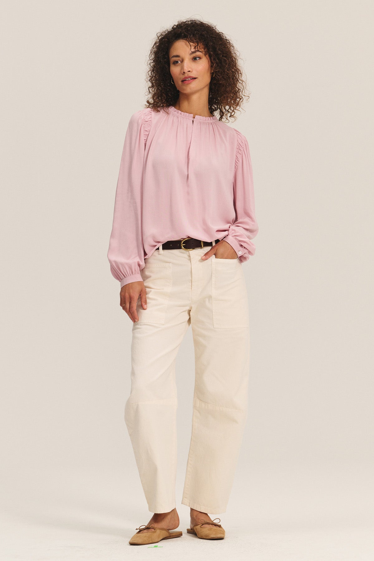 A woman stands against a plain background wearing the TAMARA BLOUSE by Velvet by Graham & Spencer, a light pink rayon challis top with ruffled neckline, paired with cream-colored pants, a black belt, and tan flats.-40598551199937