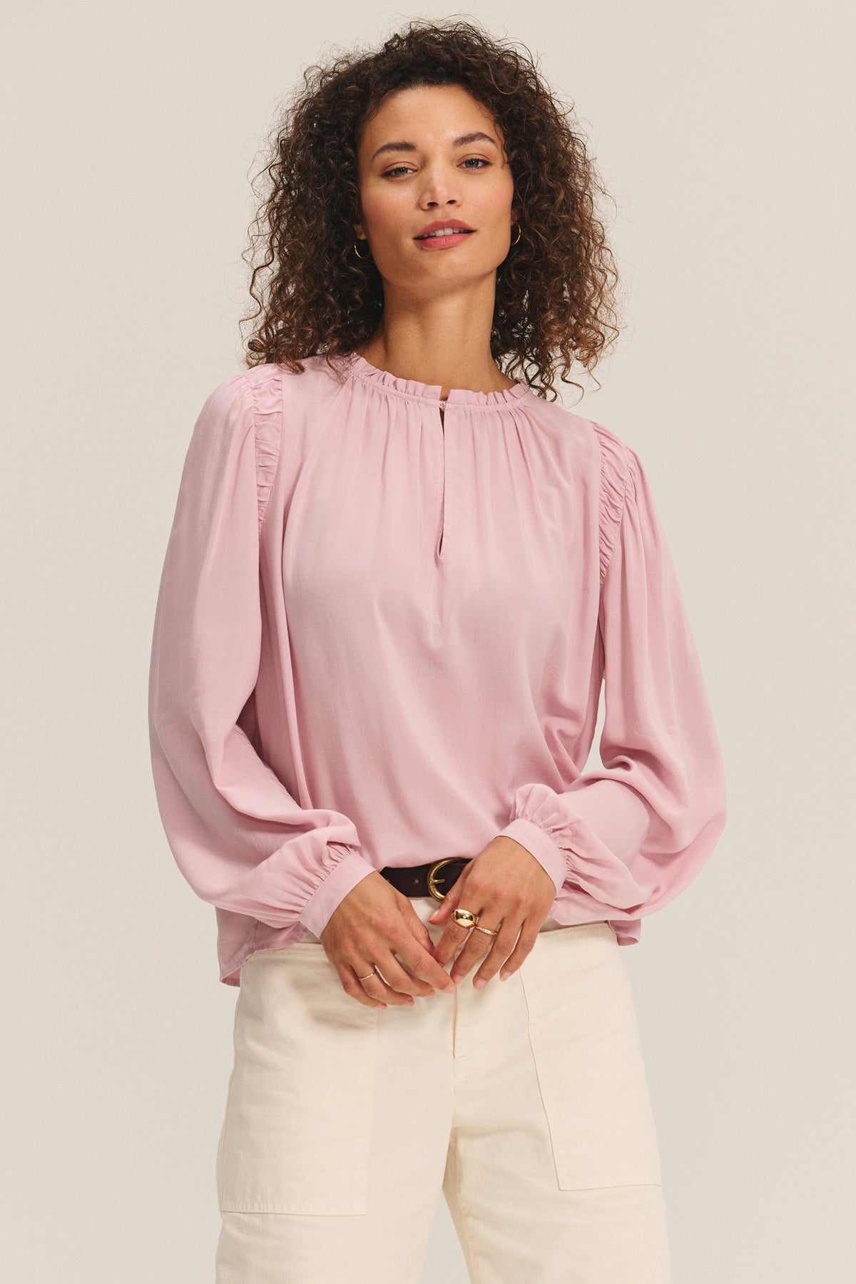 A woman wears the TAMARA BLOUSE by Velvet by Graham & Spencer—a light pink rayon challis top with gathered shoulders and a ruffled neckline—paired with cream pants, standing against a neutral background.-40598551167169