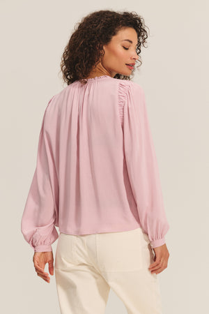 A woman is seen from the back wearing the TAMARA BLOUSE by Velvet by Graham & Spencer in light pink rayon challis with a ruffled neckline, paired with off-white pants against a plain background.